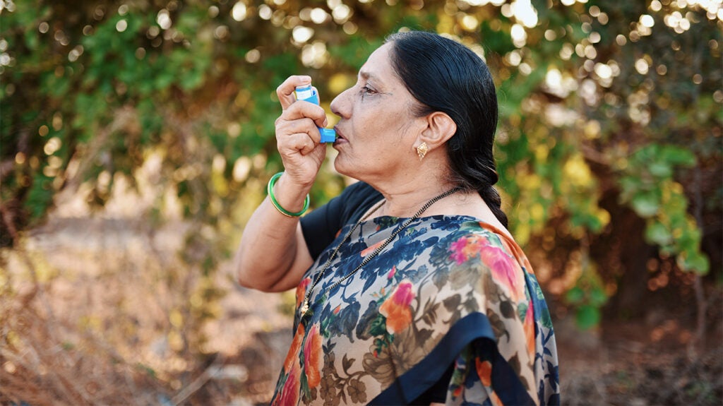 Older adult standing in the outdoors taking a puff from an asthma inhaler as part of their asthma treatment plan 